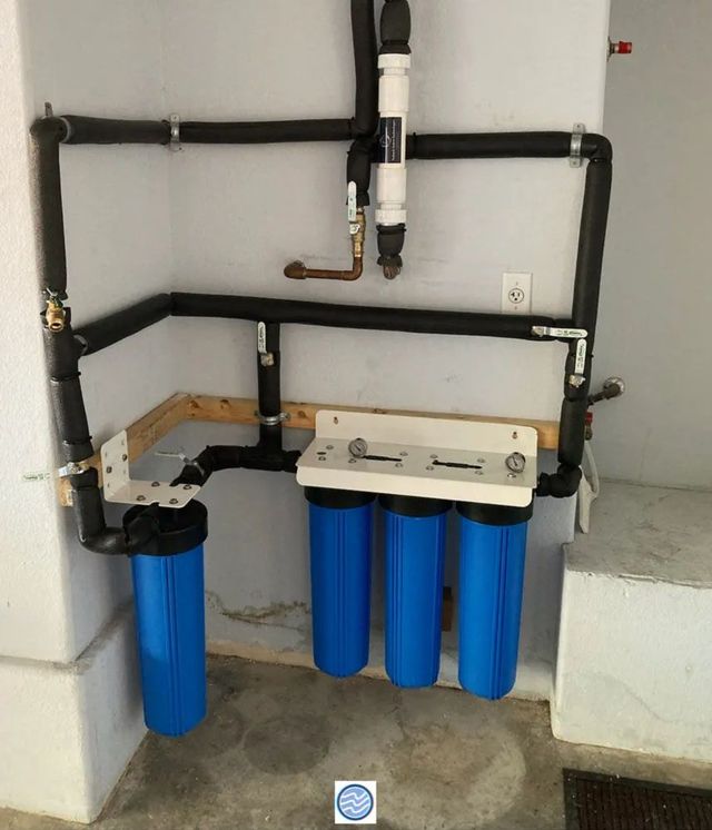 Blue water filtration system with four cartridges and black pipes mounted in a utility room