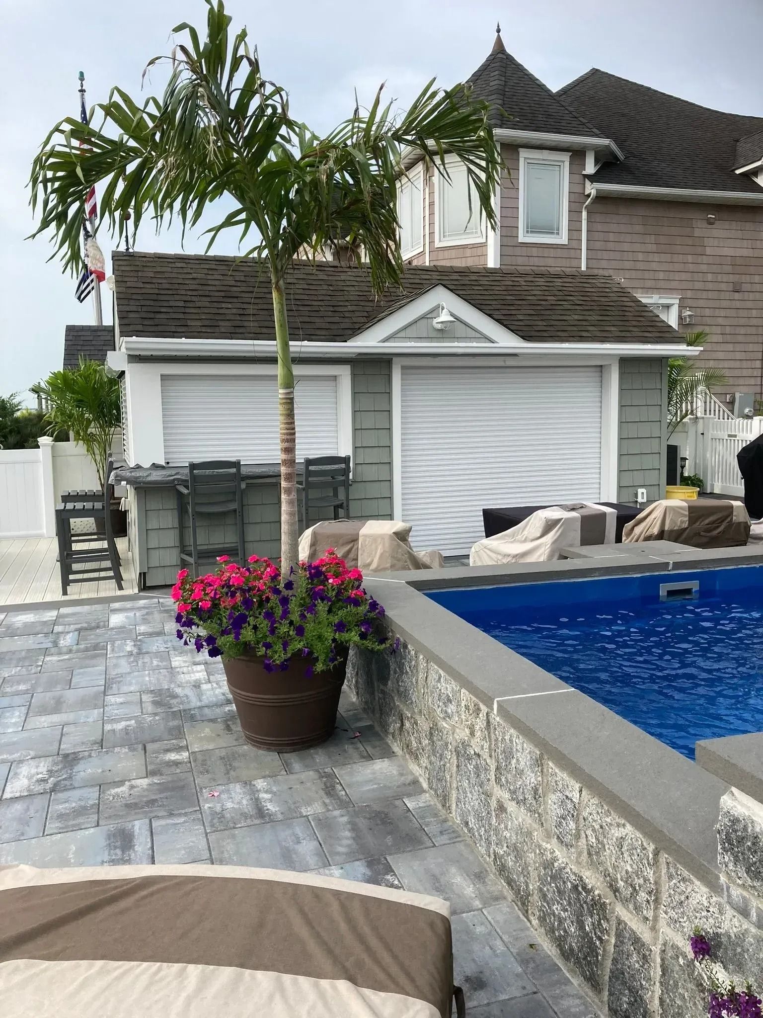 Backyard patio with pool, lounge chairs, potted flowers, and a two-story house with white shutters