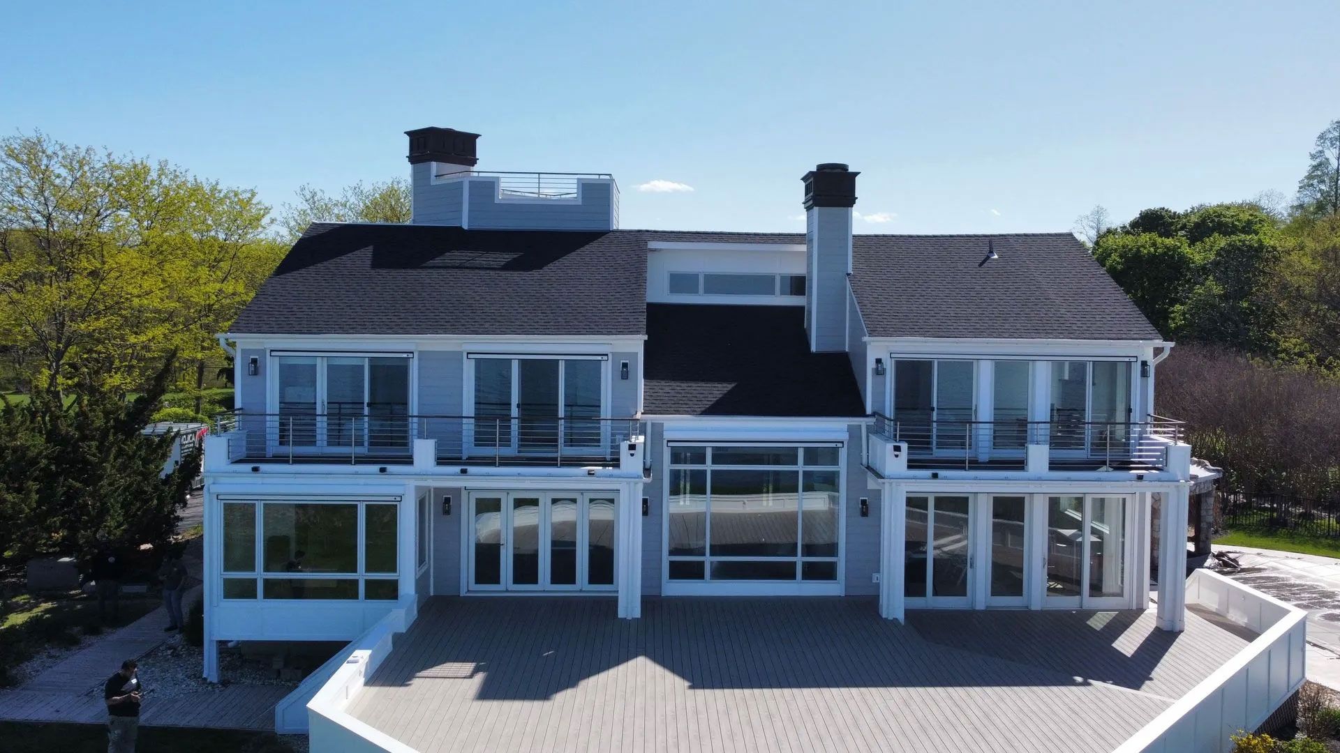 Large white waterfront house with glass sunrooms, dark roof, and a broad deck surrounded by trees