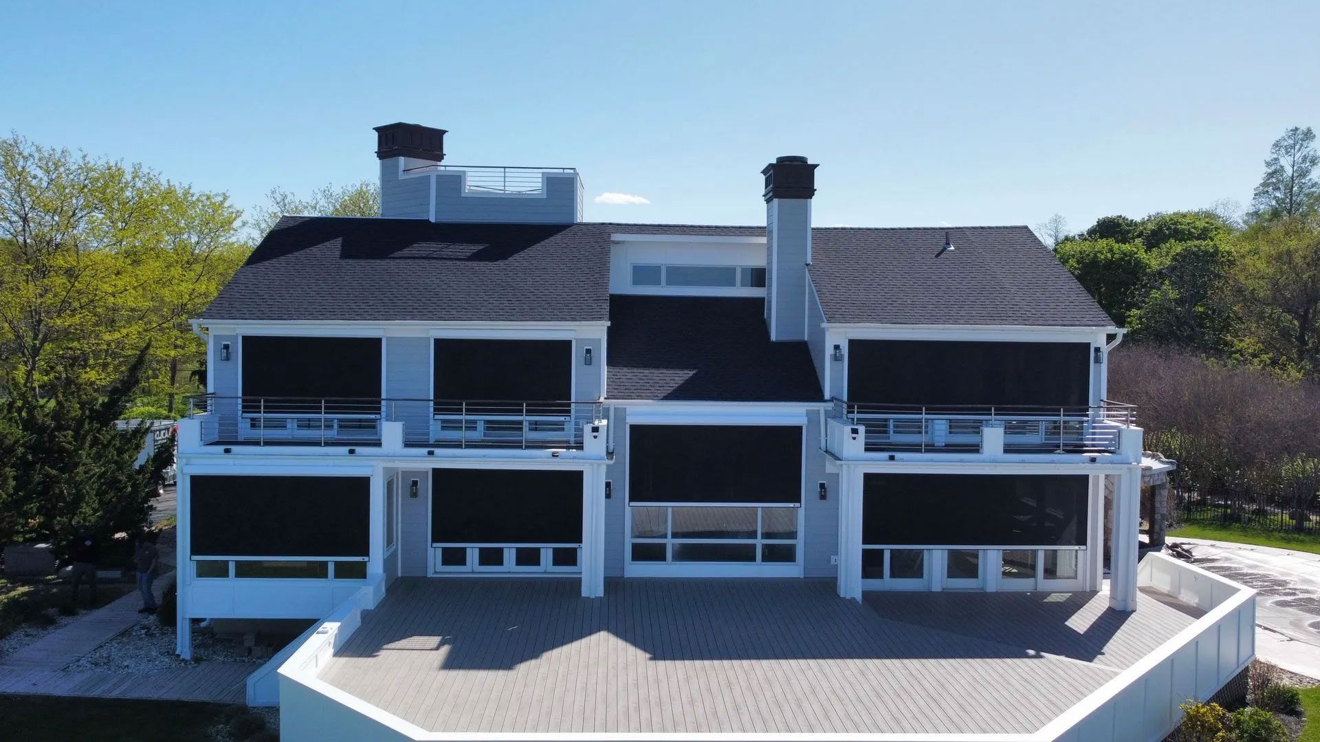 Large two-story white house with black roof and balconies on a sunny hillside above trees