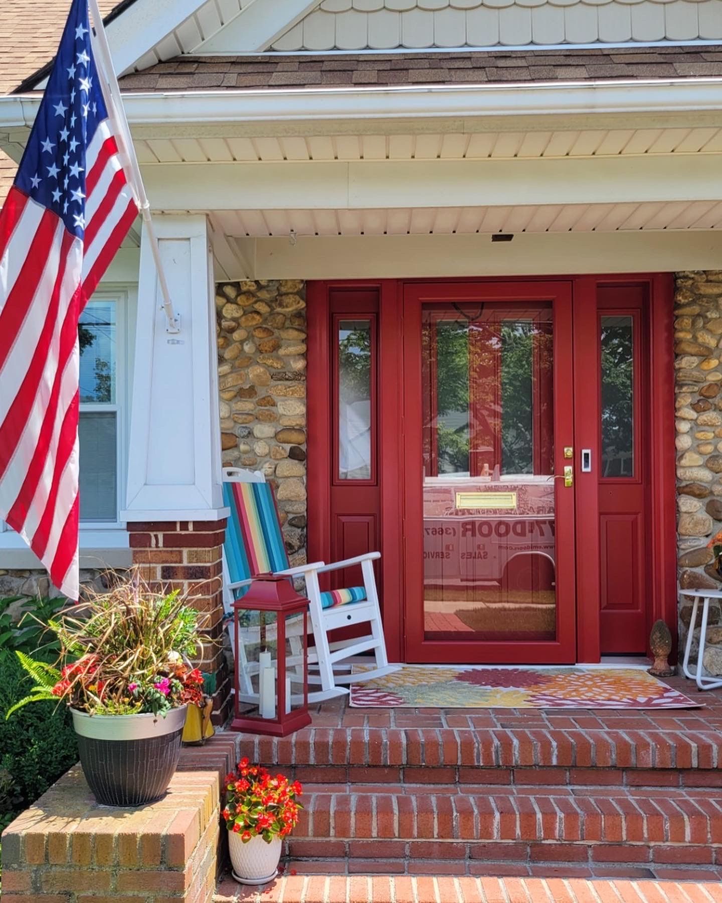 Red front door with sidelights and brick steps, decorated with an American flag, plants, and a rocking chair.