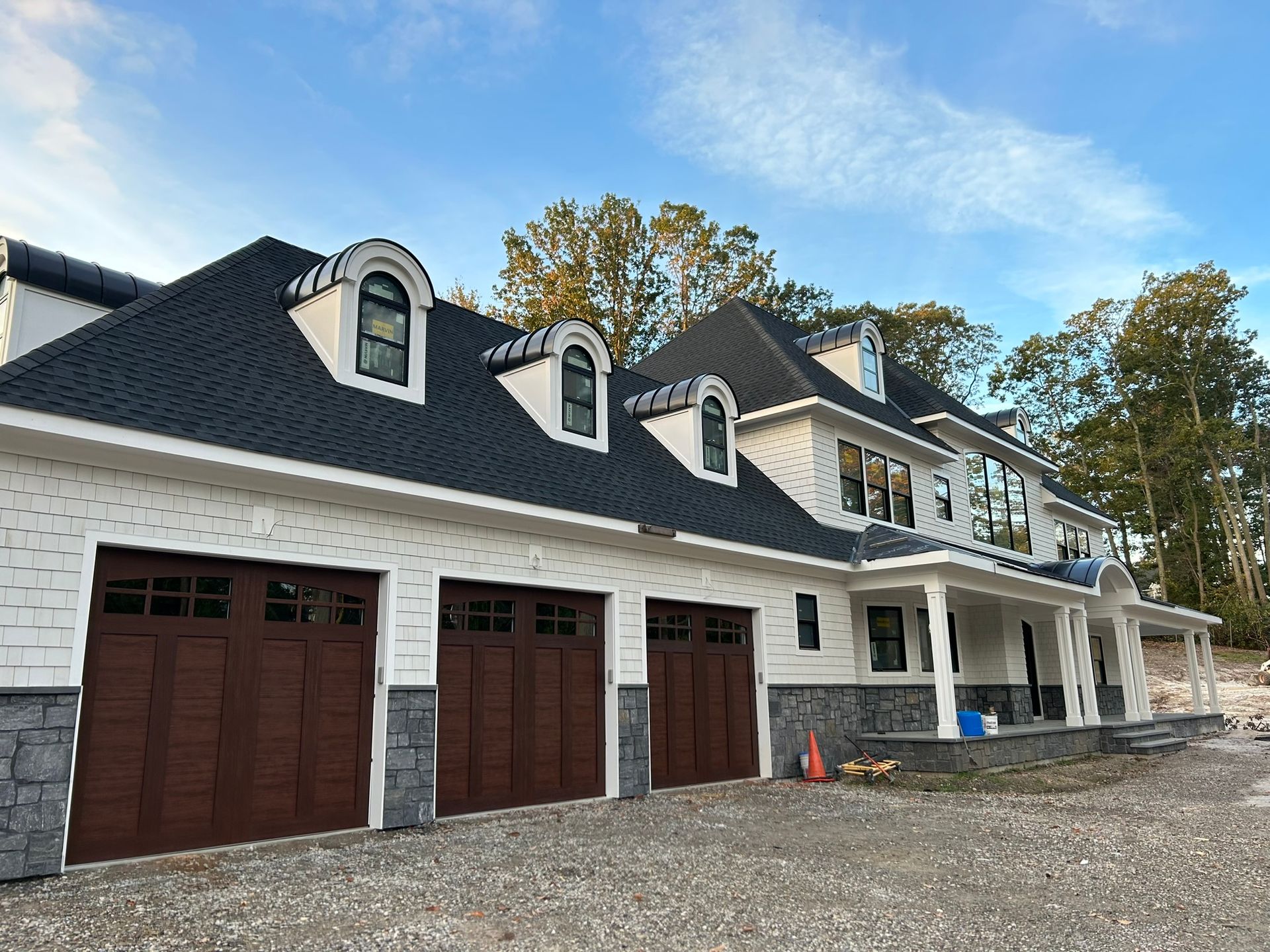 Two-story white house with a dark roof and brown garage doors on a gravel lot under a blue sky.