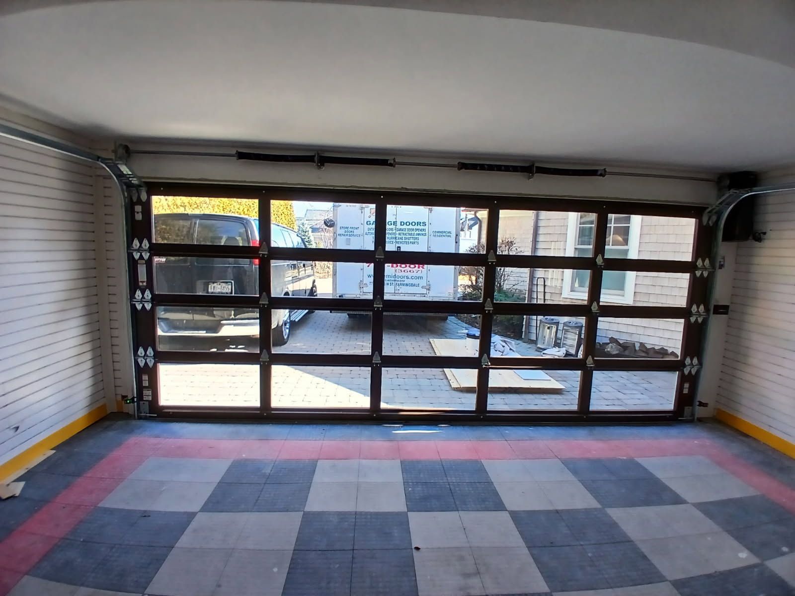Garage interior with clear glass door, checkered floor, brick walls, and view of vehicles outside.