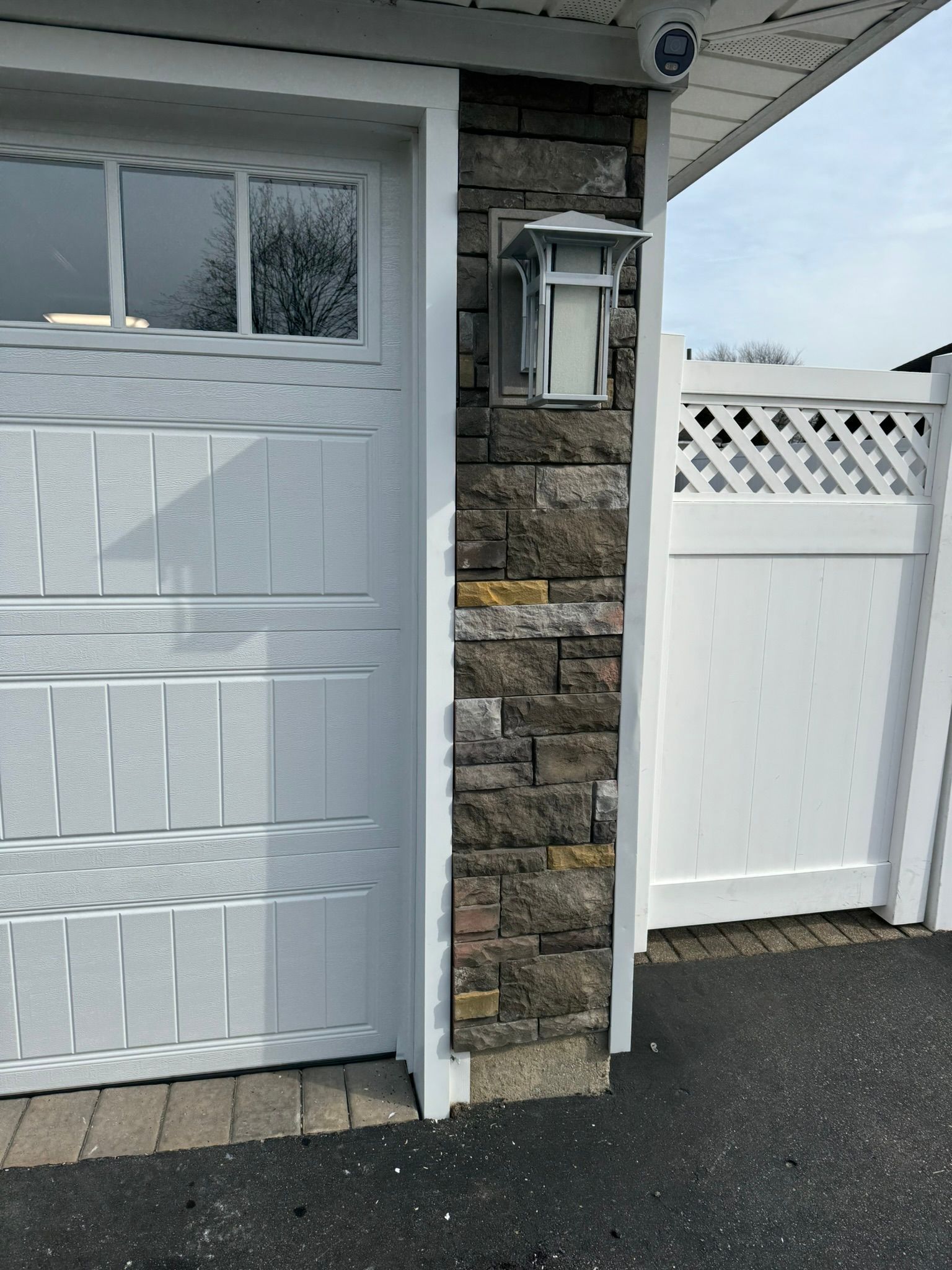 White garage door next to a stone-covered pillar with a light fixture and a white fence.