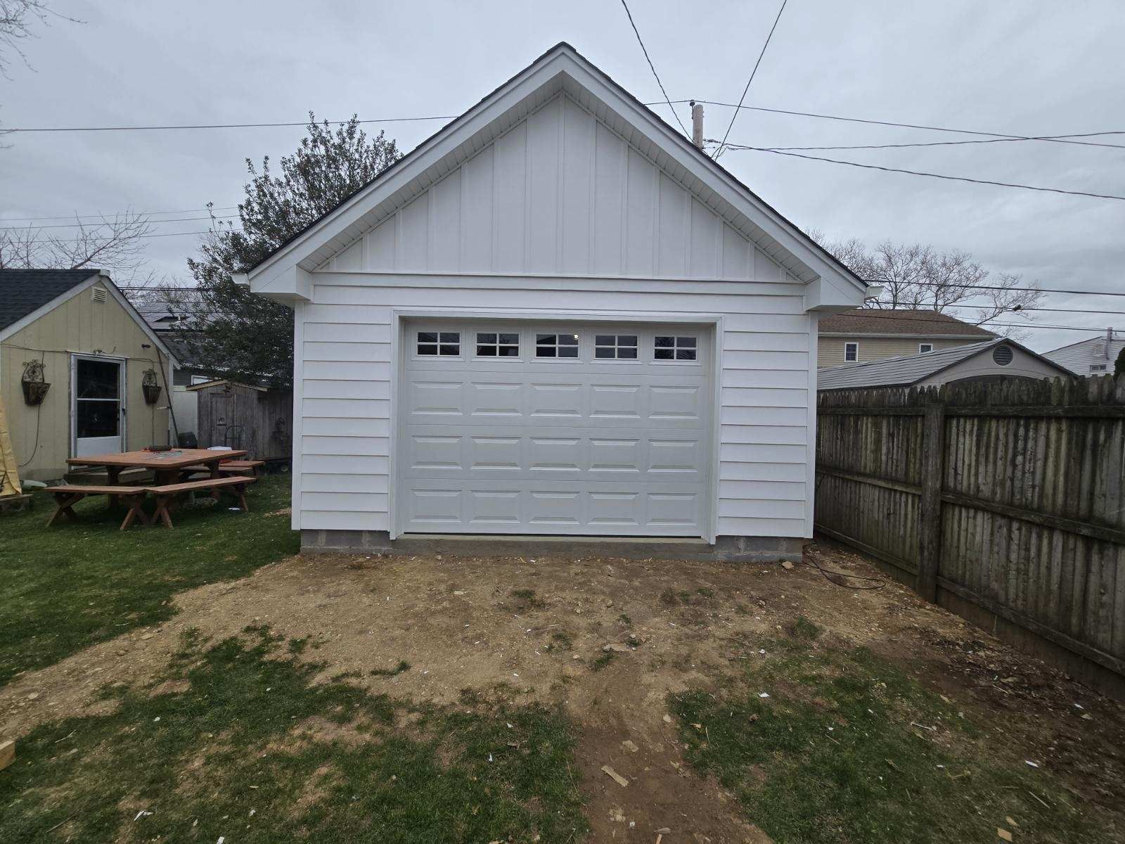 White detached garage with a gable roof, overhead door, and adjacent wooden fence on a cloudy day.