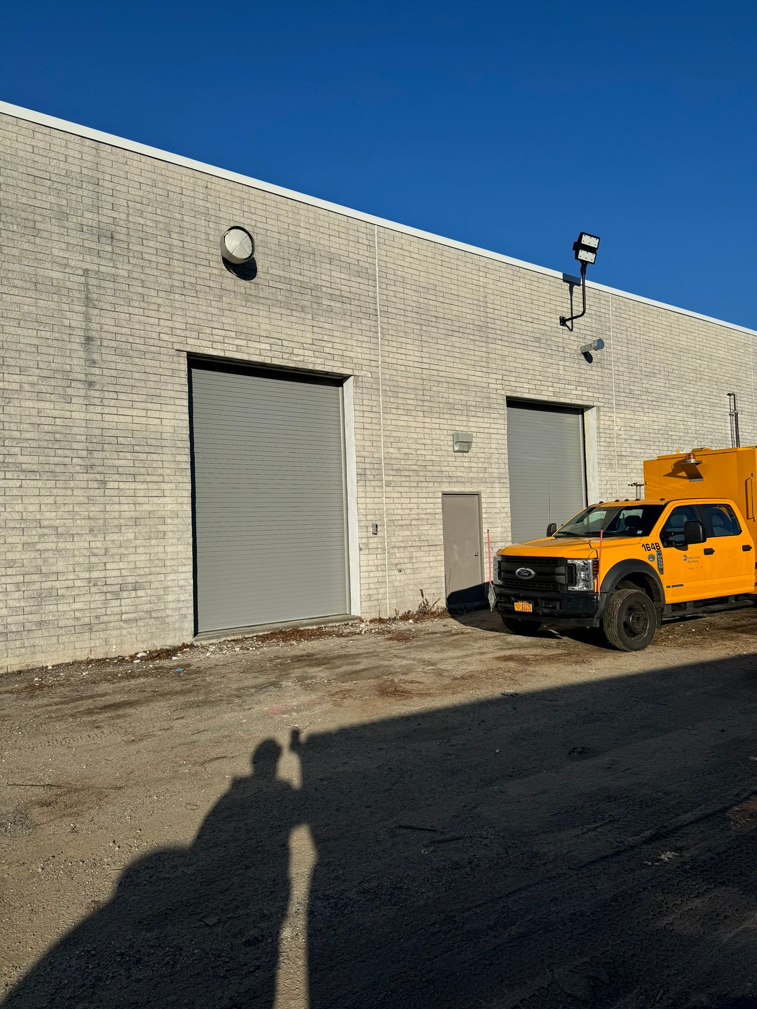 Exterior of a light brick industrial building with closed gray doors. A yellow truck is parked outside, and a person's shadow is cast on the ground.