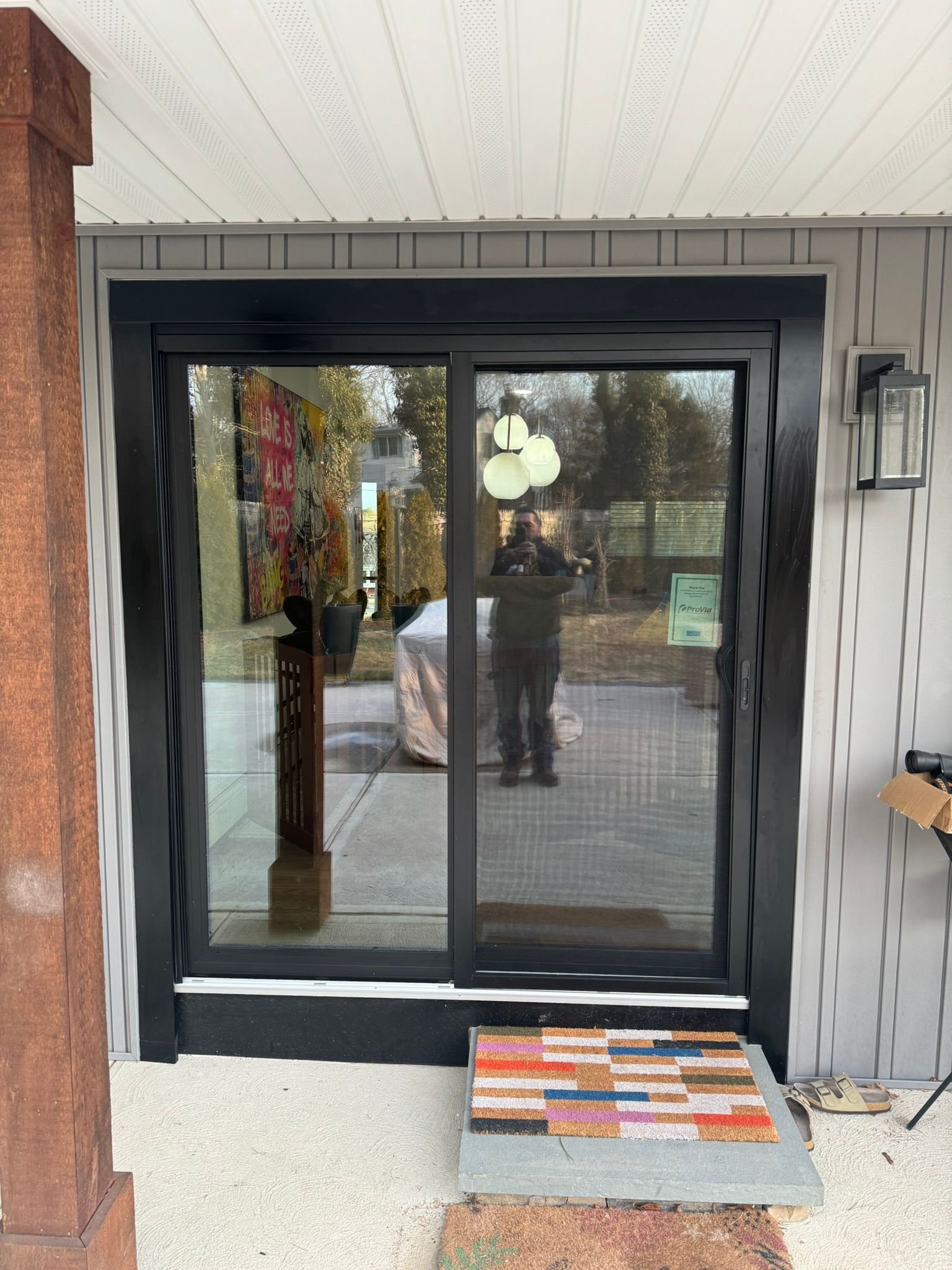 Black-framed sliding glass door with textured trim, a doormat, and a person reflected in the glass.