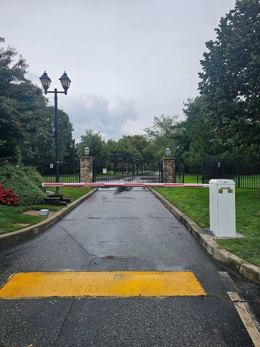 Entrance gate with a barrier arm on a wet asphalt road, flanked by trees and a security post under overcast skies.