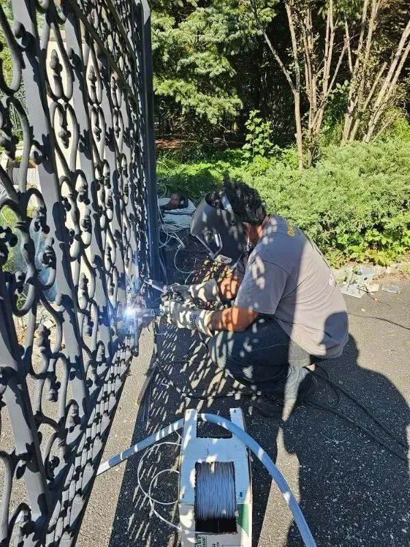 Person welding ornate metal gate outdoors, bright sunlight.