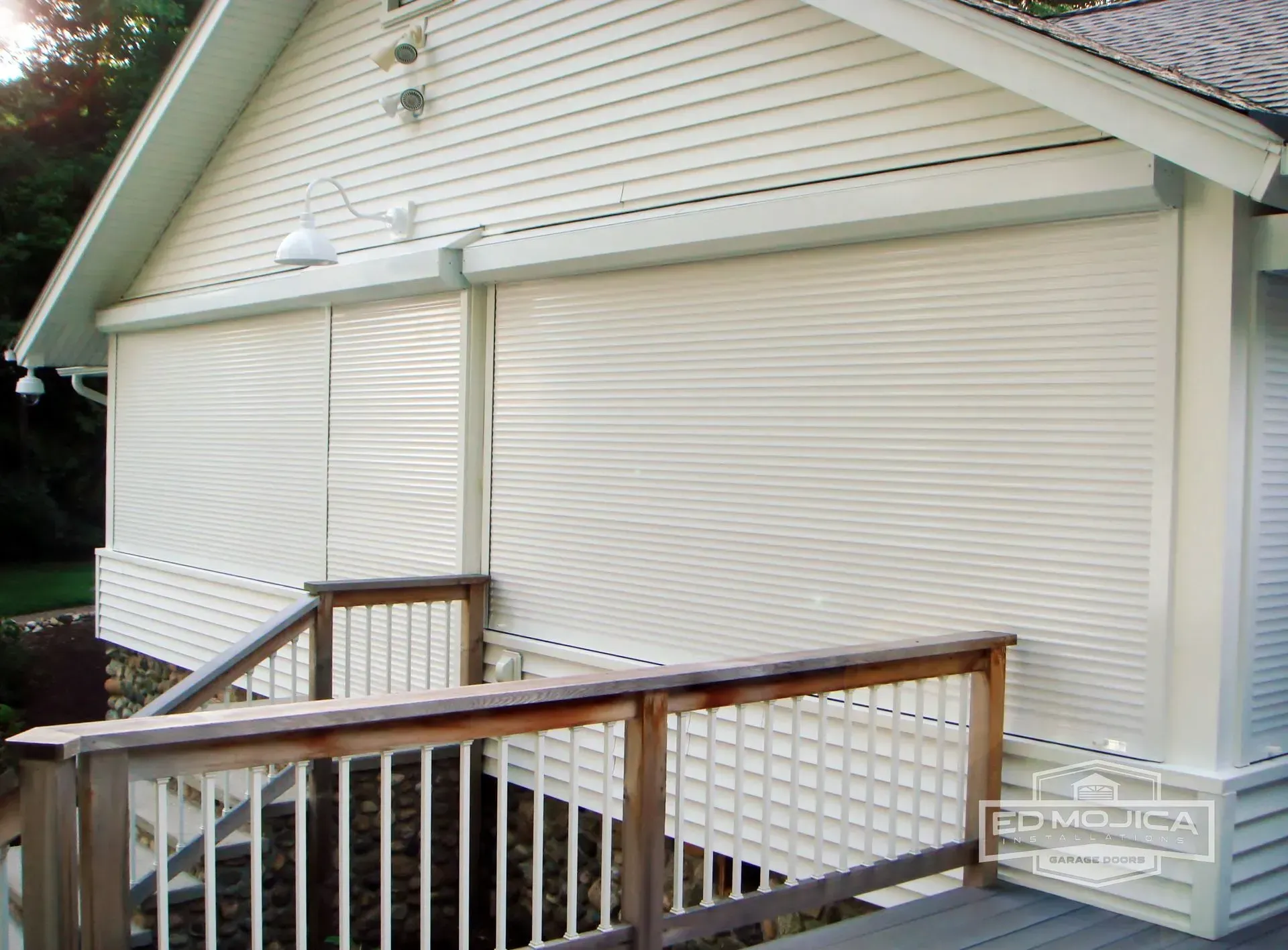 Exterior of a house with white horizontal siding and roll-down shades over windows, and a wooden deck.