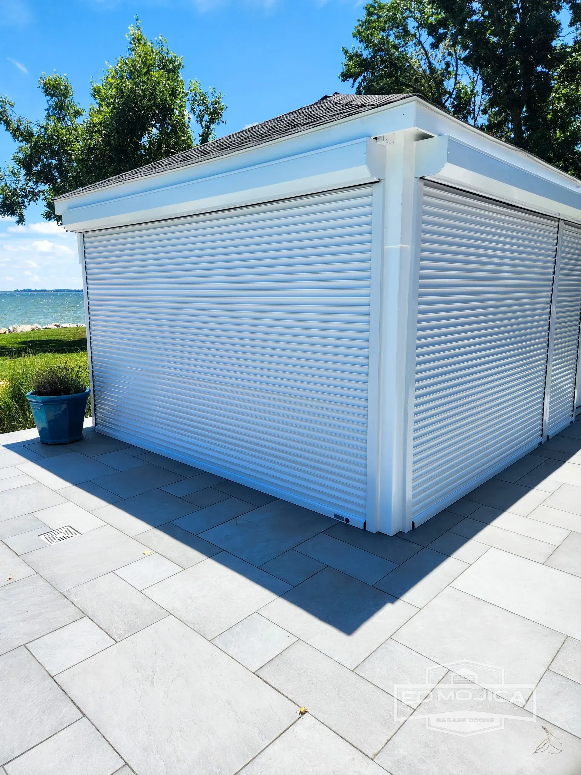 White building with rolling shutters on a stone patio, with water and trees in the background.