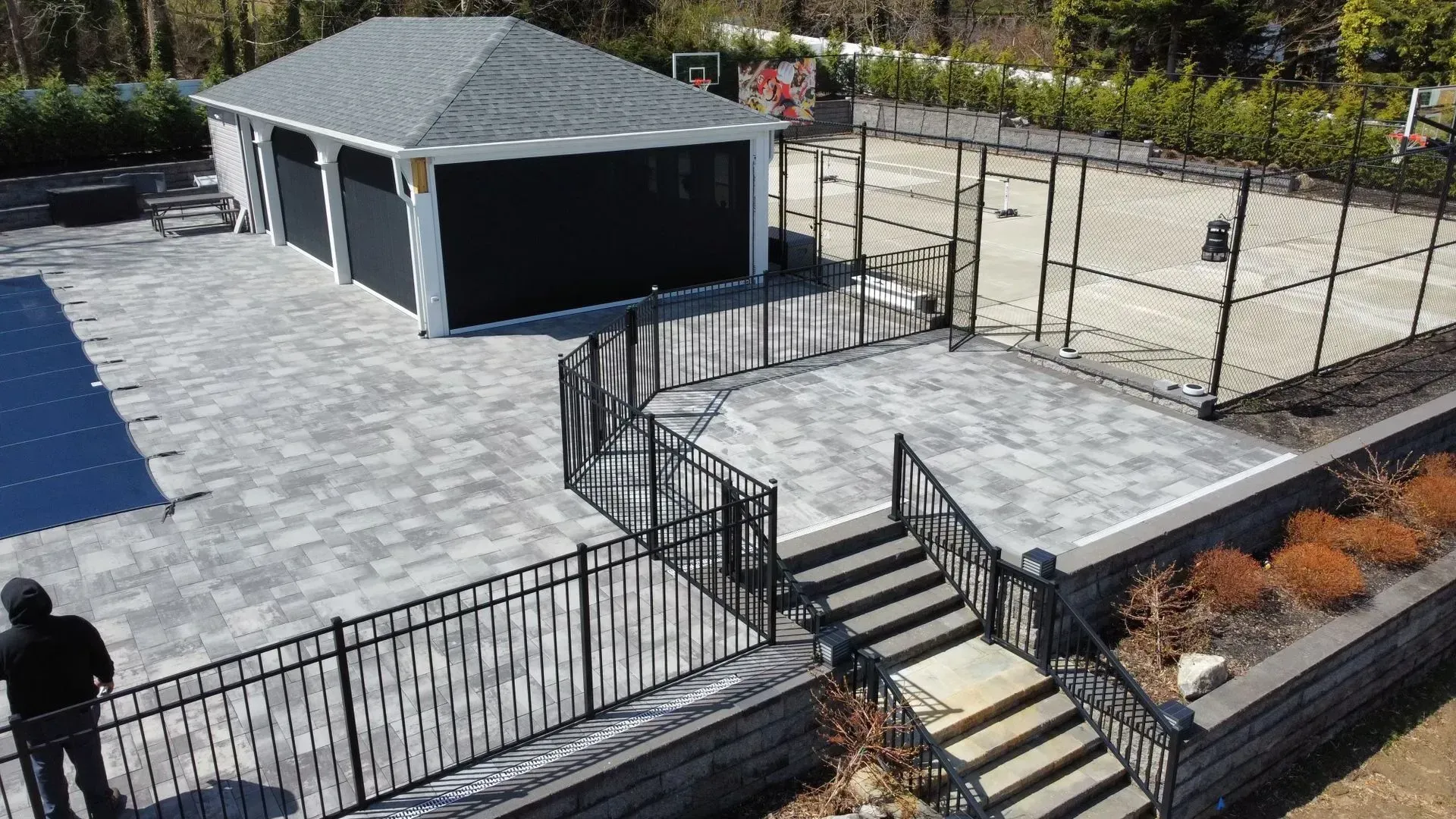 Gray paver patio with garage and tennis court, black fencing, and person.
