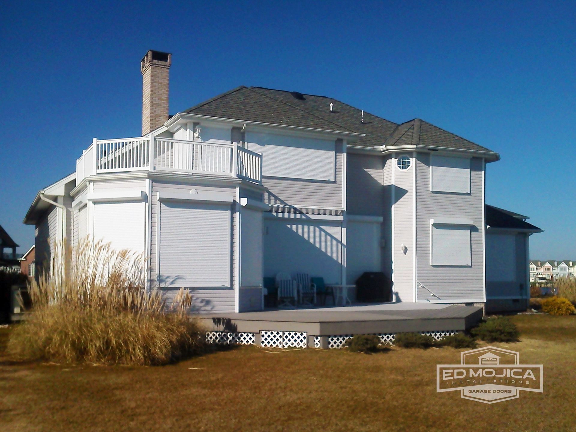 Two-story house with gray shutters and roof, a deck, and a balcony on a sunny day.