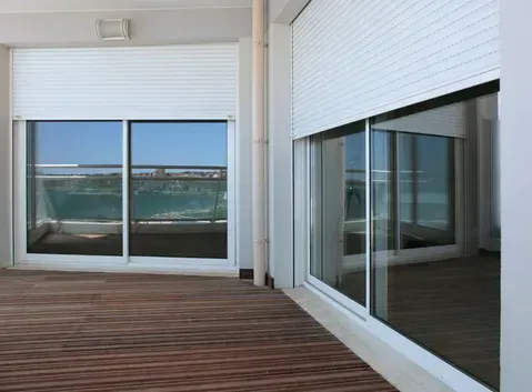 Exterior view of a balcony with closed white shutters and large windows reflecting a seascape.