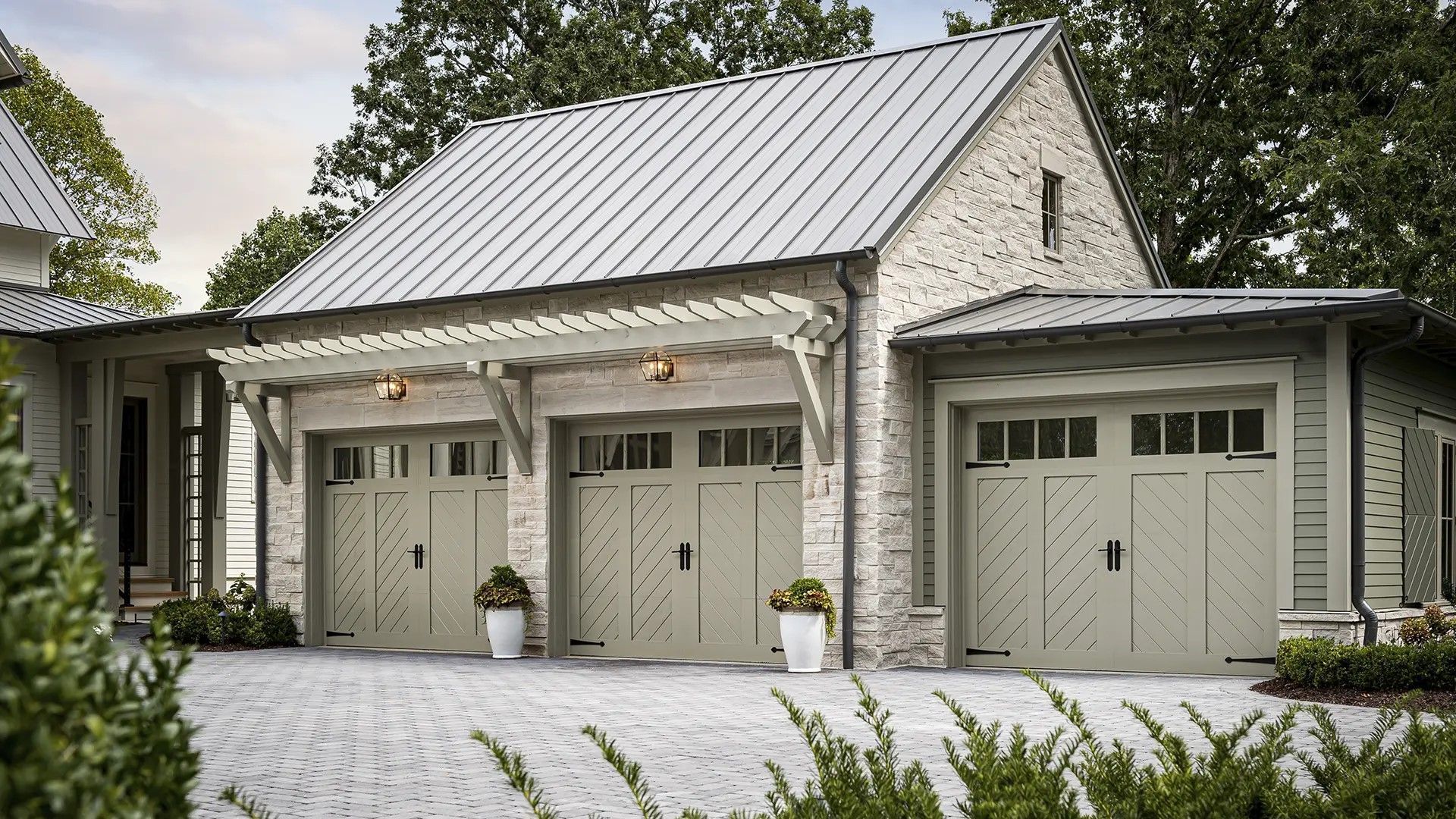 Three-car garage with light green doors, stone exterior, and gray metal roof on a cobblestone driveway.