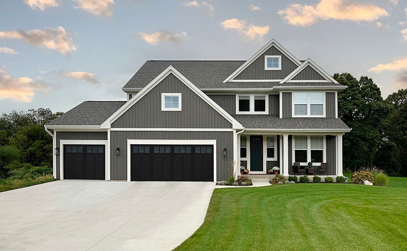 Gray two-story house with a driveway, garage doors, and green lawn under a cloudy sky.