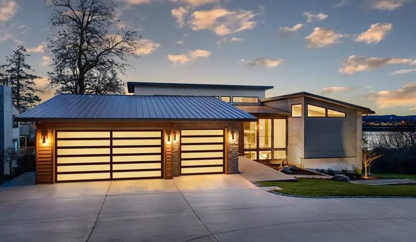 Modern home with glass garage doors, illuminated by warm light, overlooking a lake at sunset.