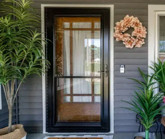 Black framed front door with glass and screen, on a gray house with wreath and plants.