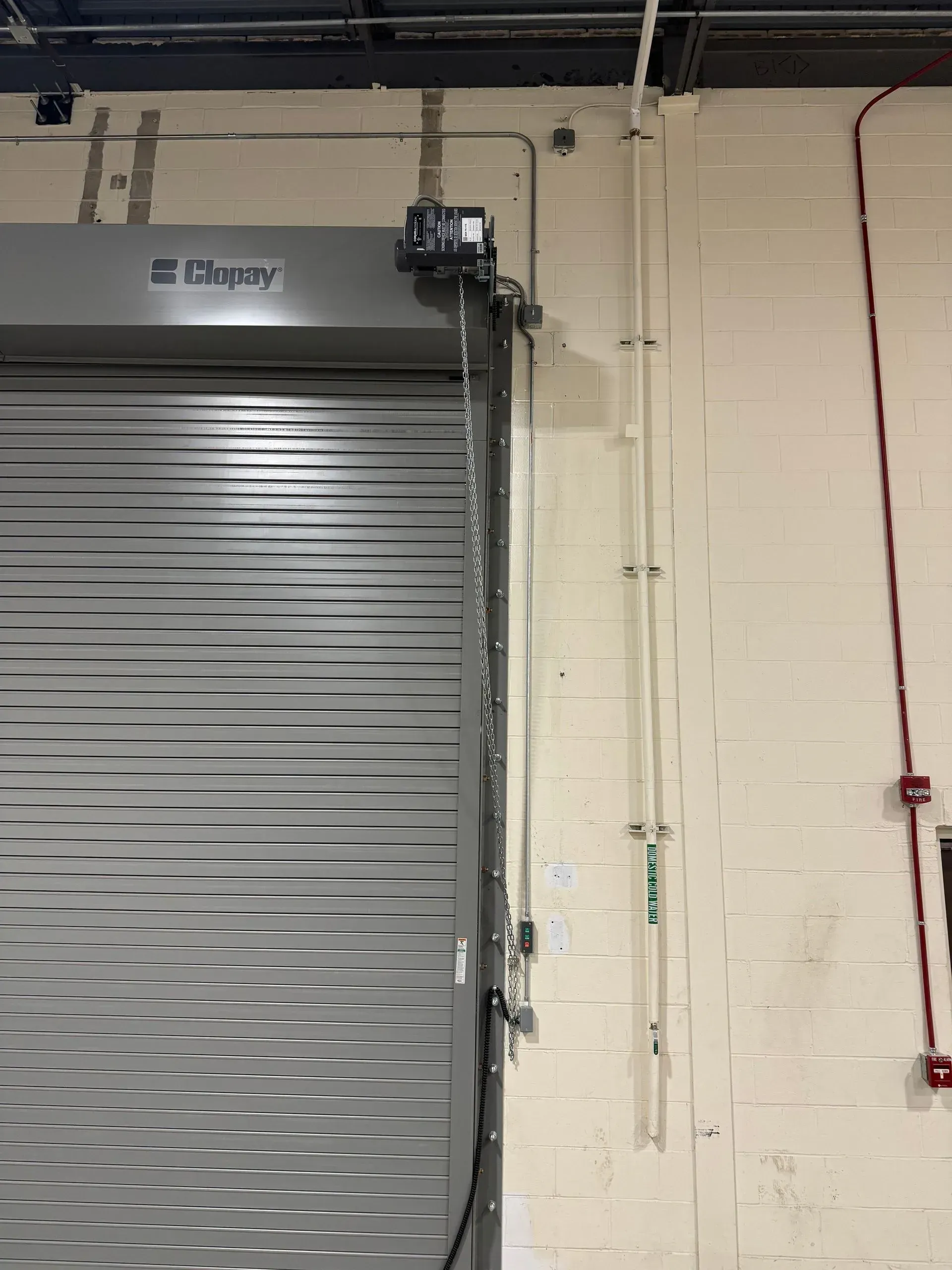 Roll-up security door, grey and corrugated, partially open. Motor mechanism and chain visible. 