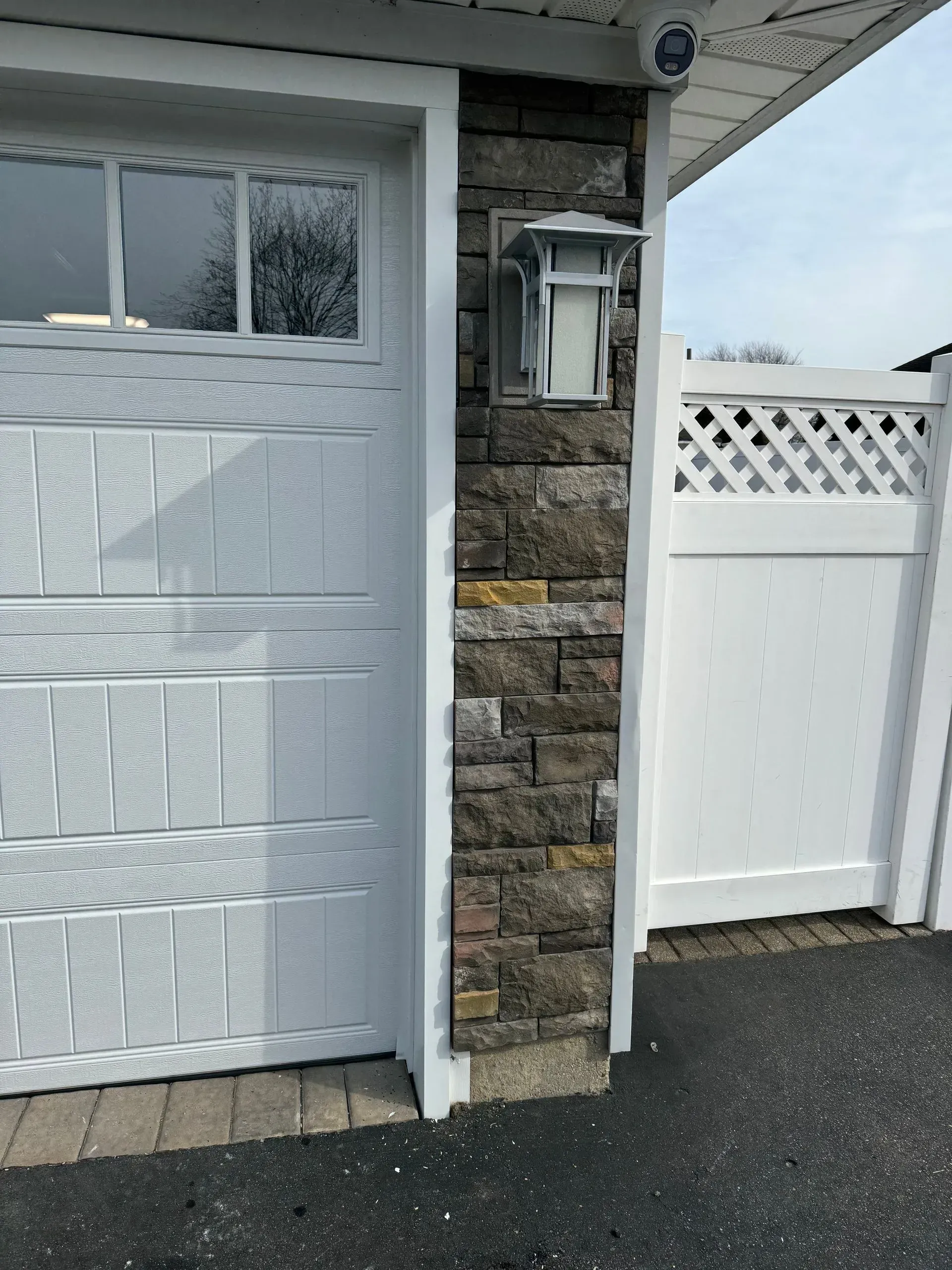 White garage door next to a stone-covered pillar with a light fixture and a white fence on the right.