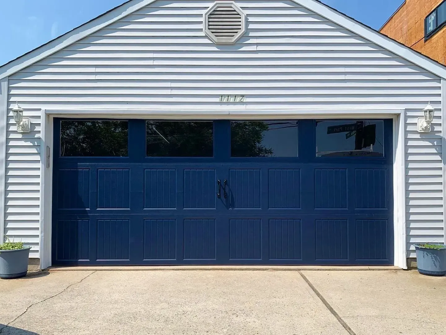 Blue garage door with windows, on a light blue-sided building with potted plants.