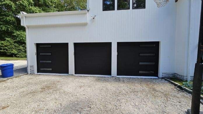 Three black garage doors on a white building, gravel driveway, and a blue trash can.