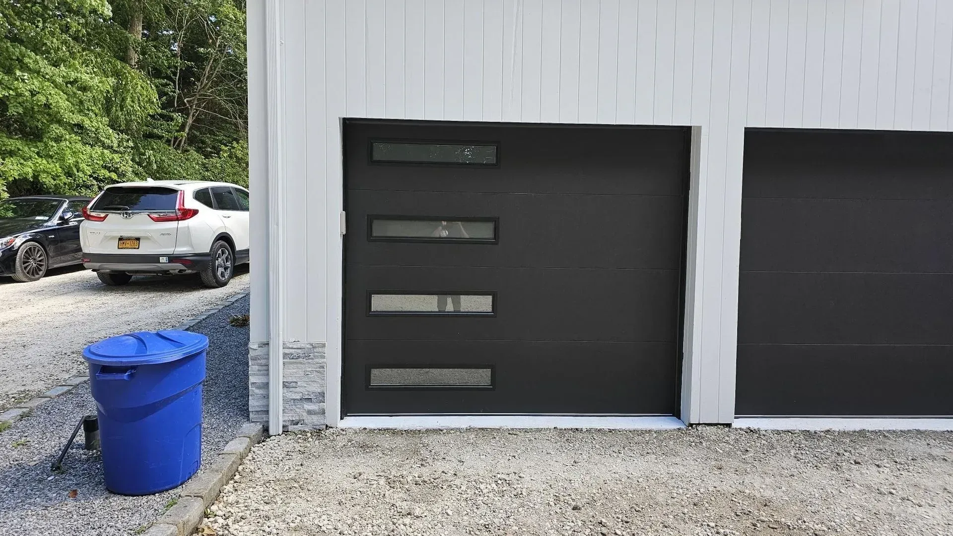 Black garage door with horizontal glass windows, blue trash bin, car on gravel.