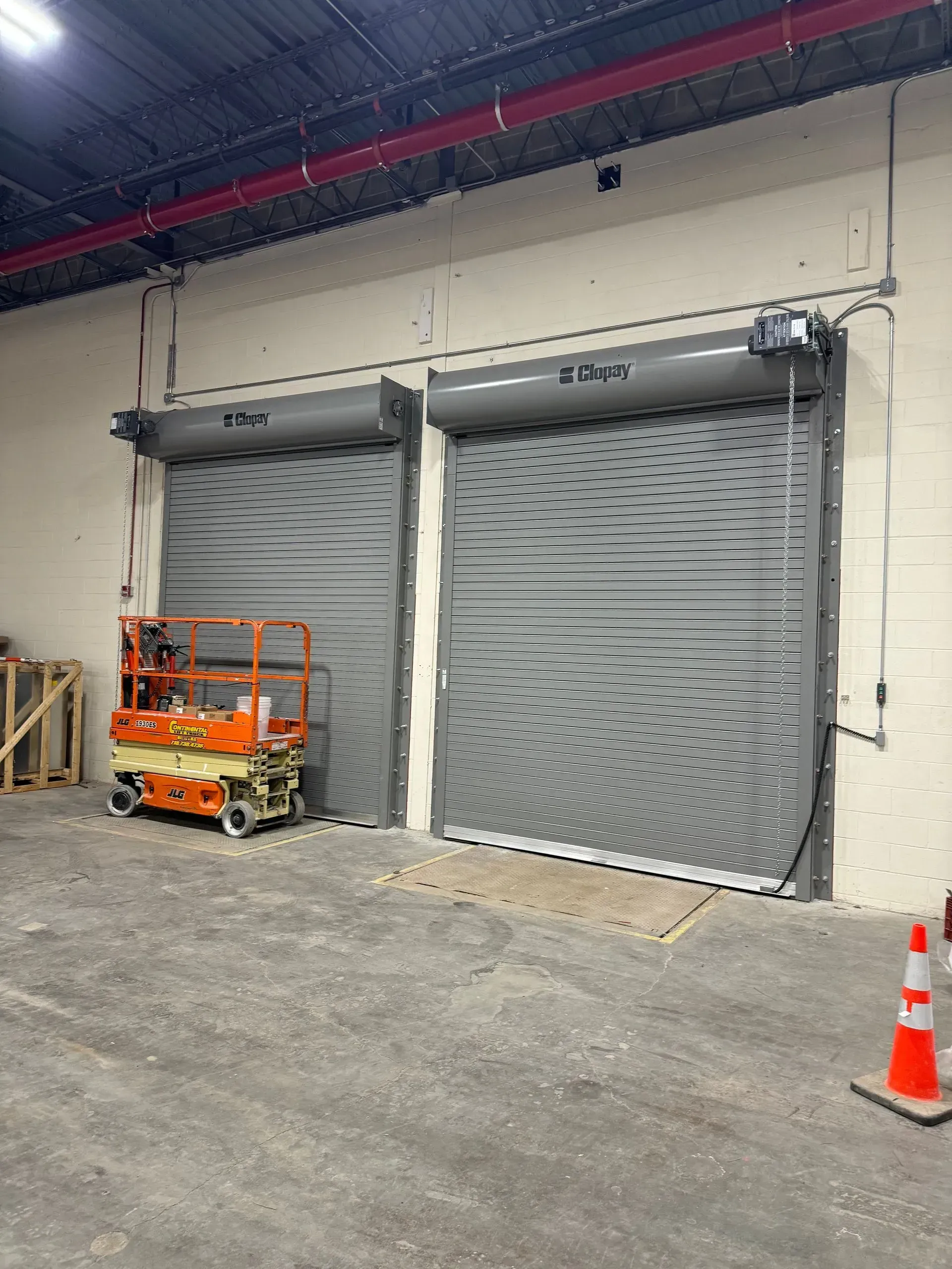 Two gray roll-up doors in a warehouse setting, with an orange lift and safety cone present.