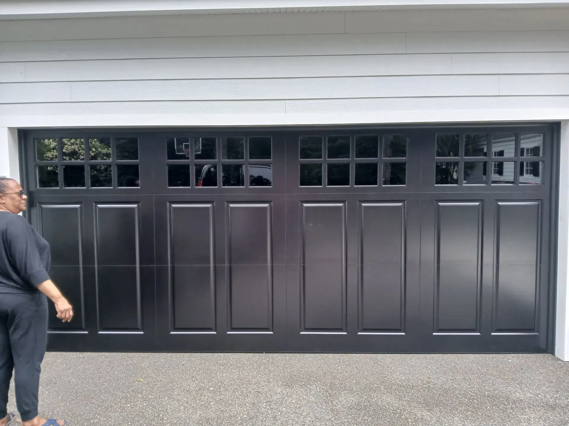 Black garage door with glass windows; a person stands nearby on a concrete driveway.