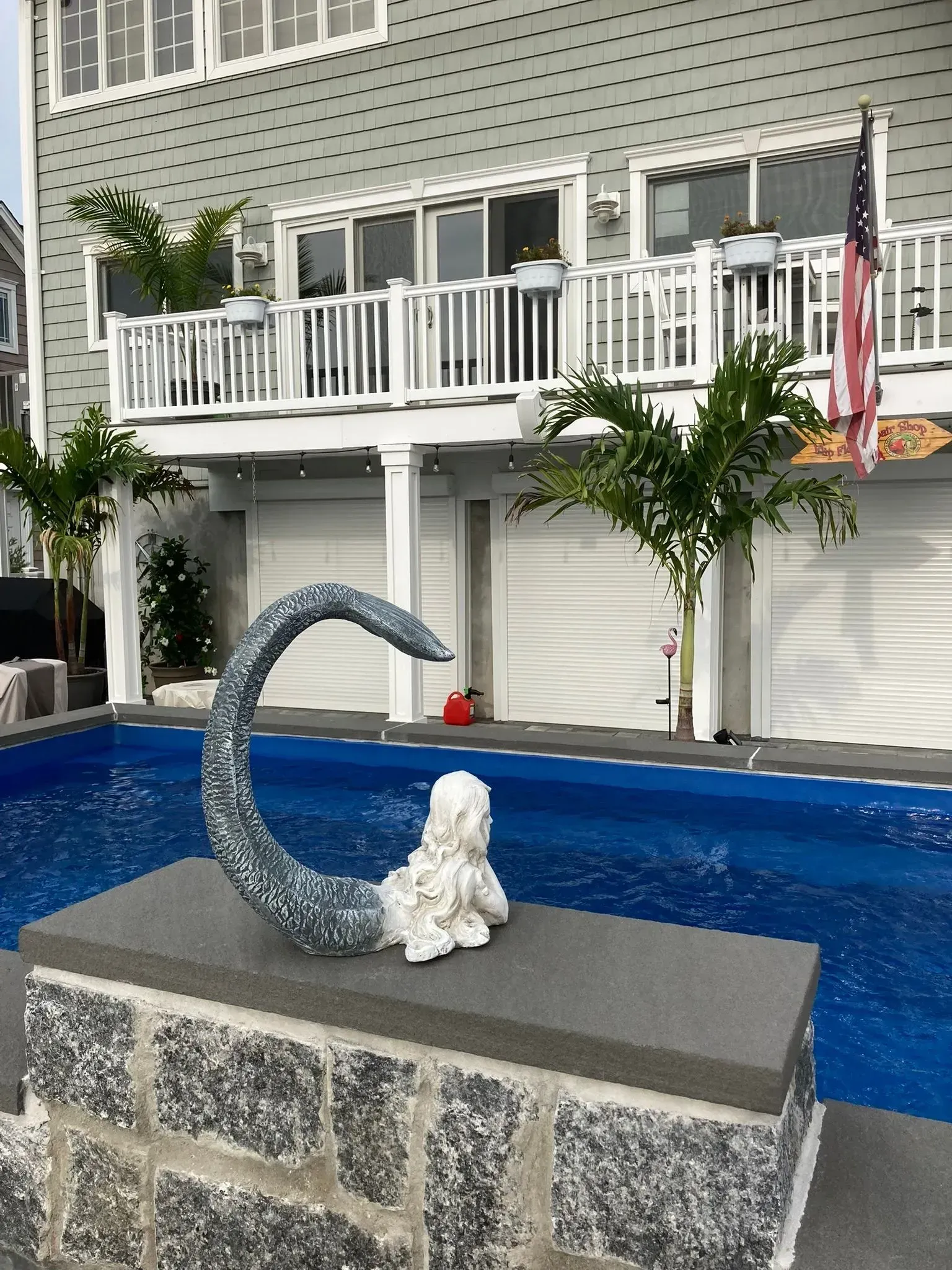 Backyard pool with sculpture, two-story house with balcony, palm trees, and American flag.
