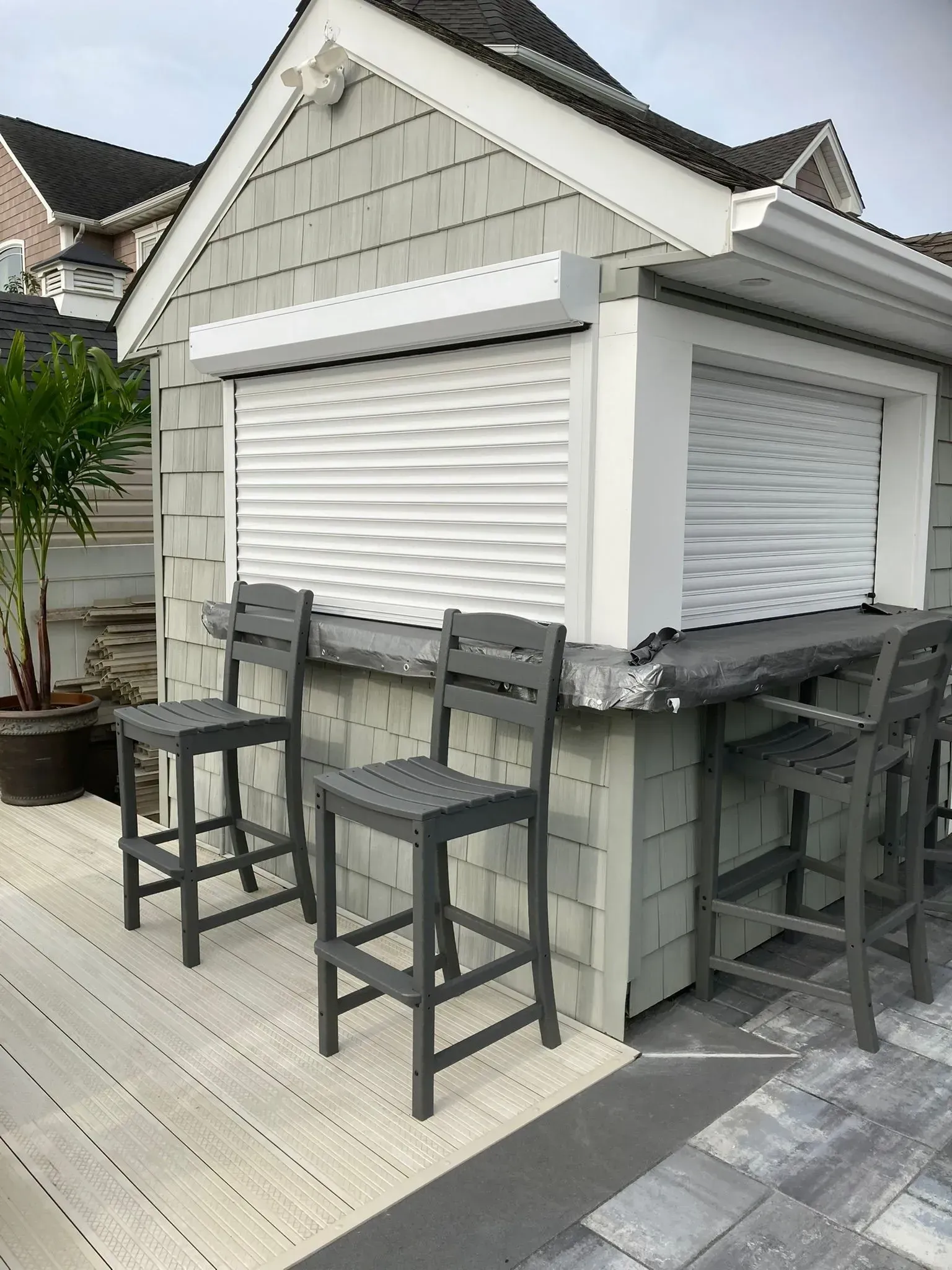 Outdoor bar with roll-down shutters, gray siding, and bar stools on a deck.