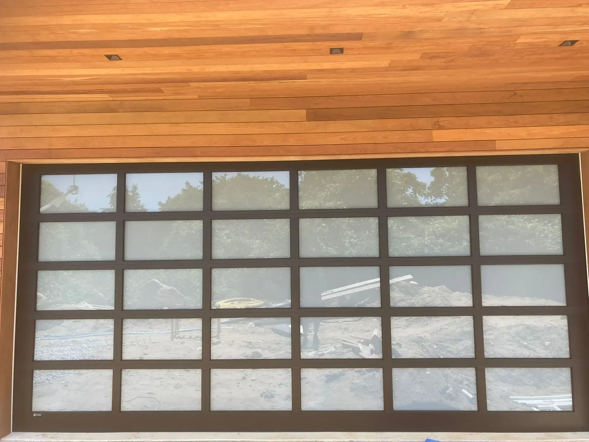 Glass garage door with grid pattern, set in wooden frame, reflecting trees and construction site.