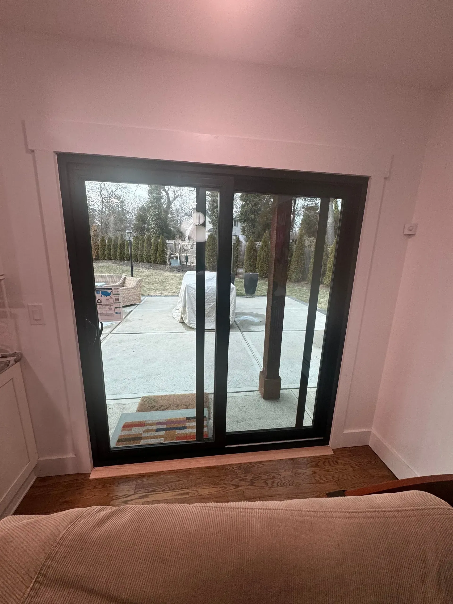 Sliding glass door, black frame, leads to a patio. Indoor view, brown floor, white trim. Outside view, yard, trees.