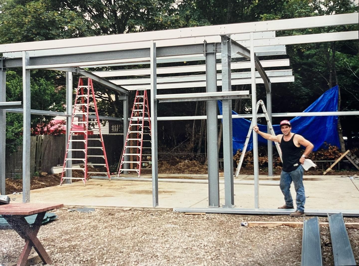 Man standing beside steel frame building under construction, holding a level. Ladders and blue tarp visible.