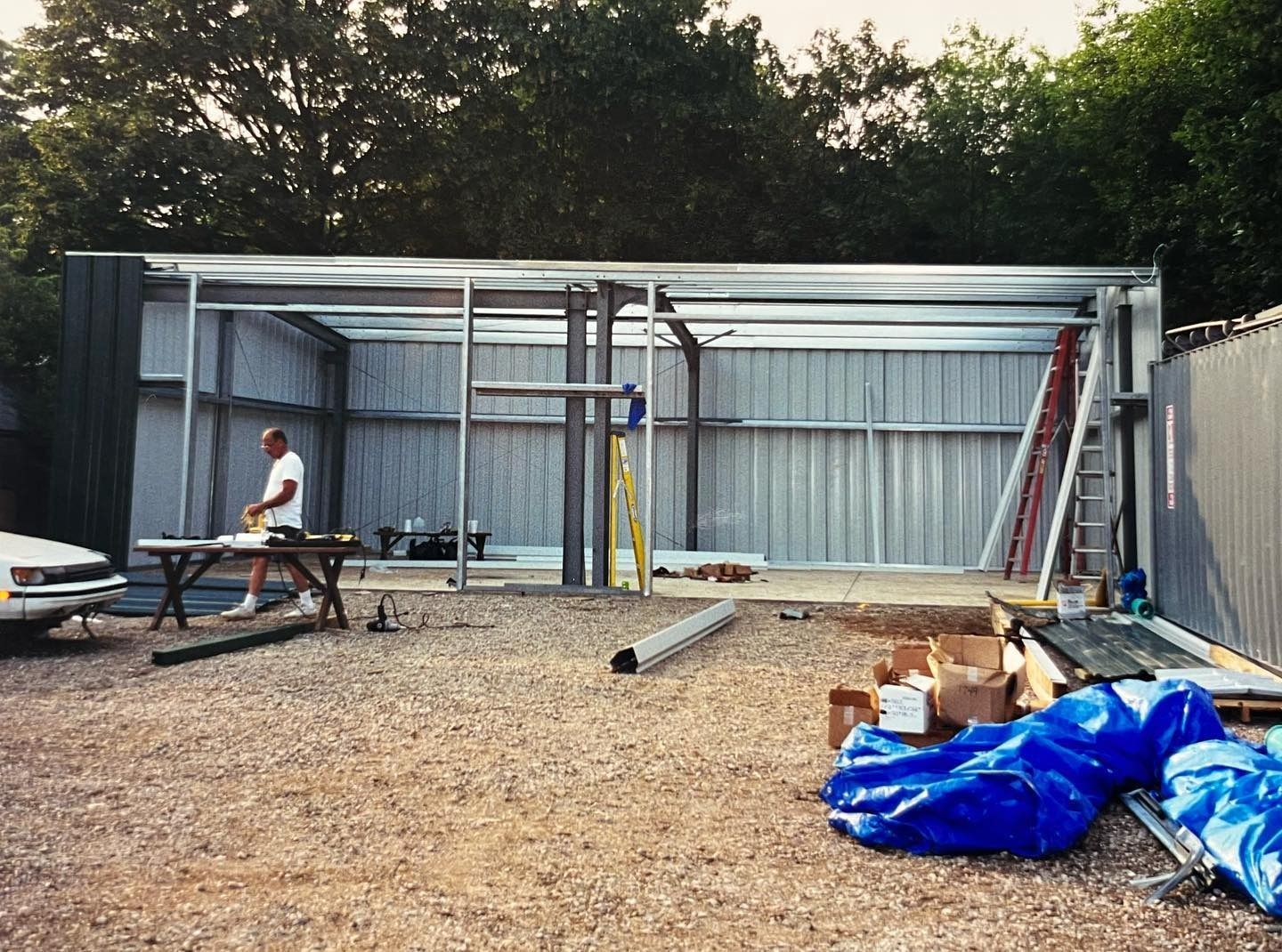 Man working on metal building under construction, exterior view. Gravel lot, tools, materials present.