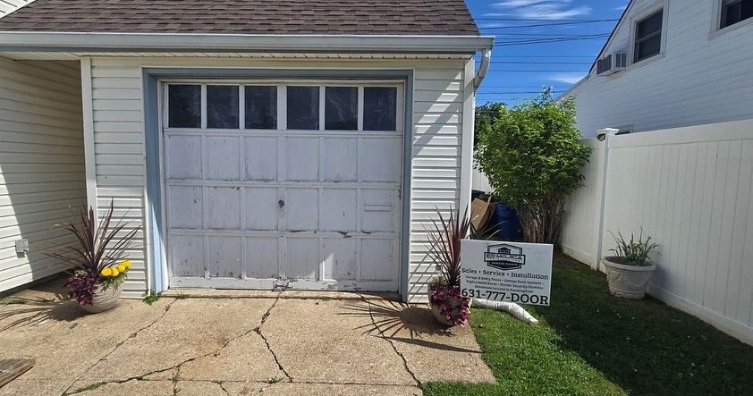 White garage with weathered door, two potted plants, and sign on the lawn.