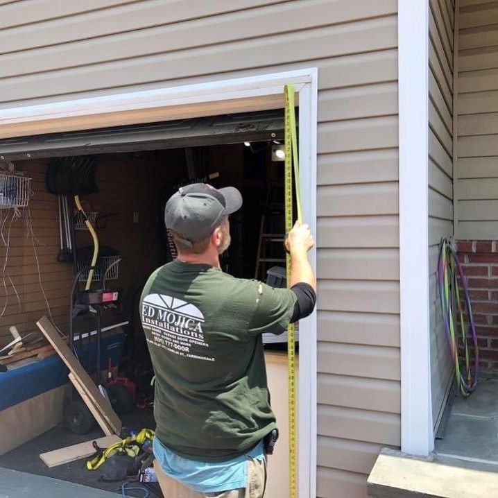 A person in a green shirt measures a garage door frame with a yellow tape measure outdoors.