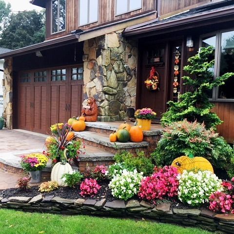 Fall-decorated home entrance with pumpkins, flowers, and a wreath. Brown siding, stone facade, and a spruce.