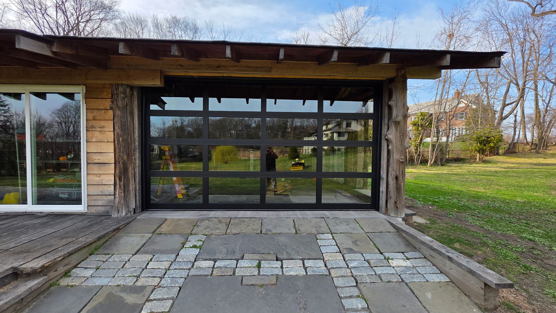 Outdoor stone patio before a wooden pavilion with large glass doors and a grass yard on the right