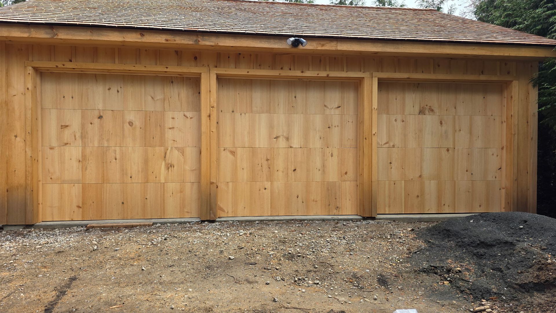 Wooden garage with three tan doors and a patterned roof, viewed from a gravel driveway.