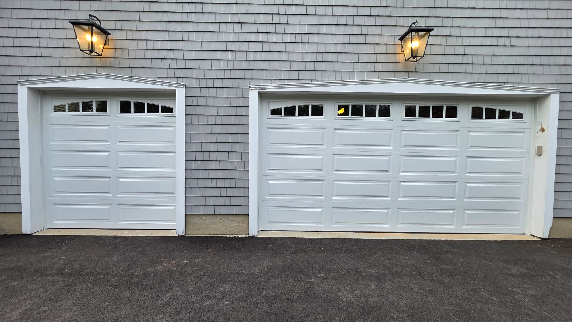 Two white garage doors on a gray house exterior, each lit by a wall lantern.