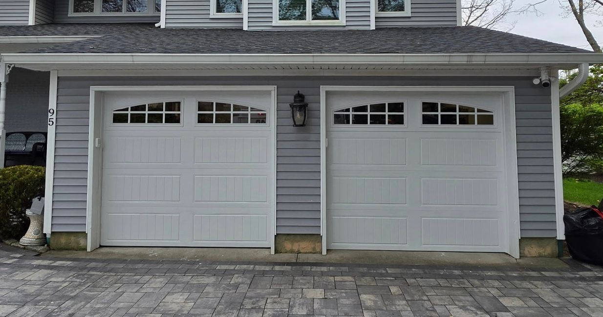 Two white garage doors on the front of a gray house with a stone driveway.