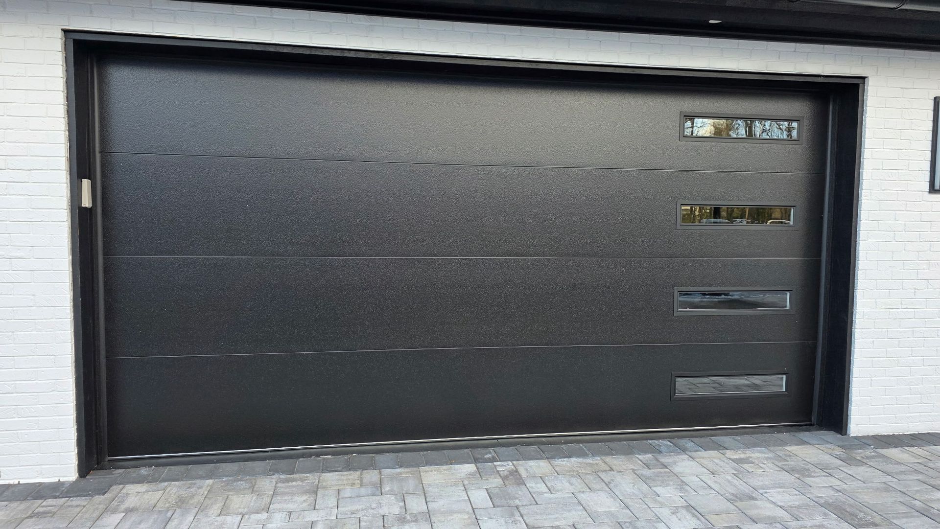 Black garage door with small windows, set in a light brick wall and paved driveway.