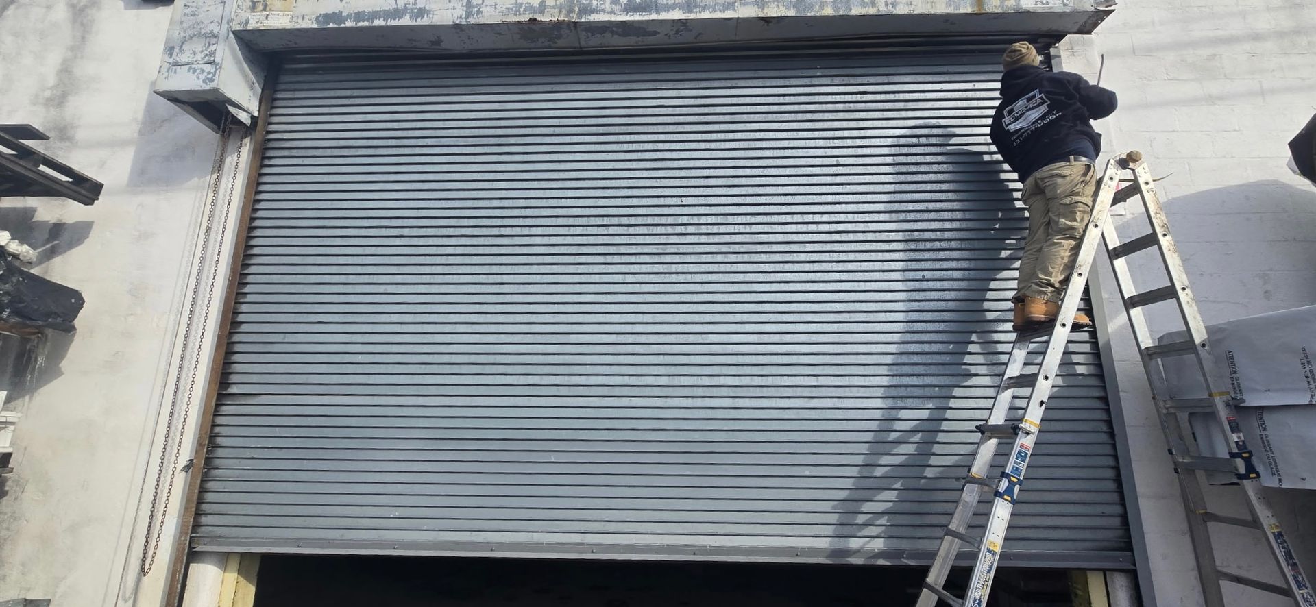 Worker on a ladder fixing a gray roll-up metal garage door on a building.