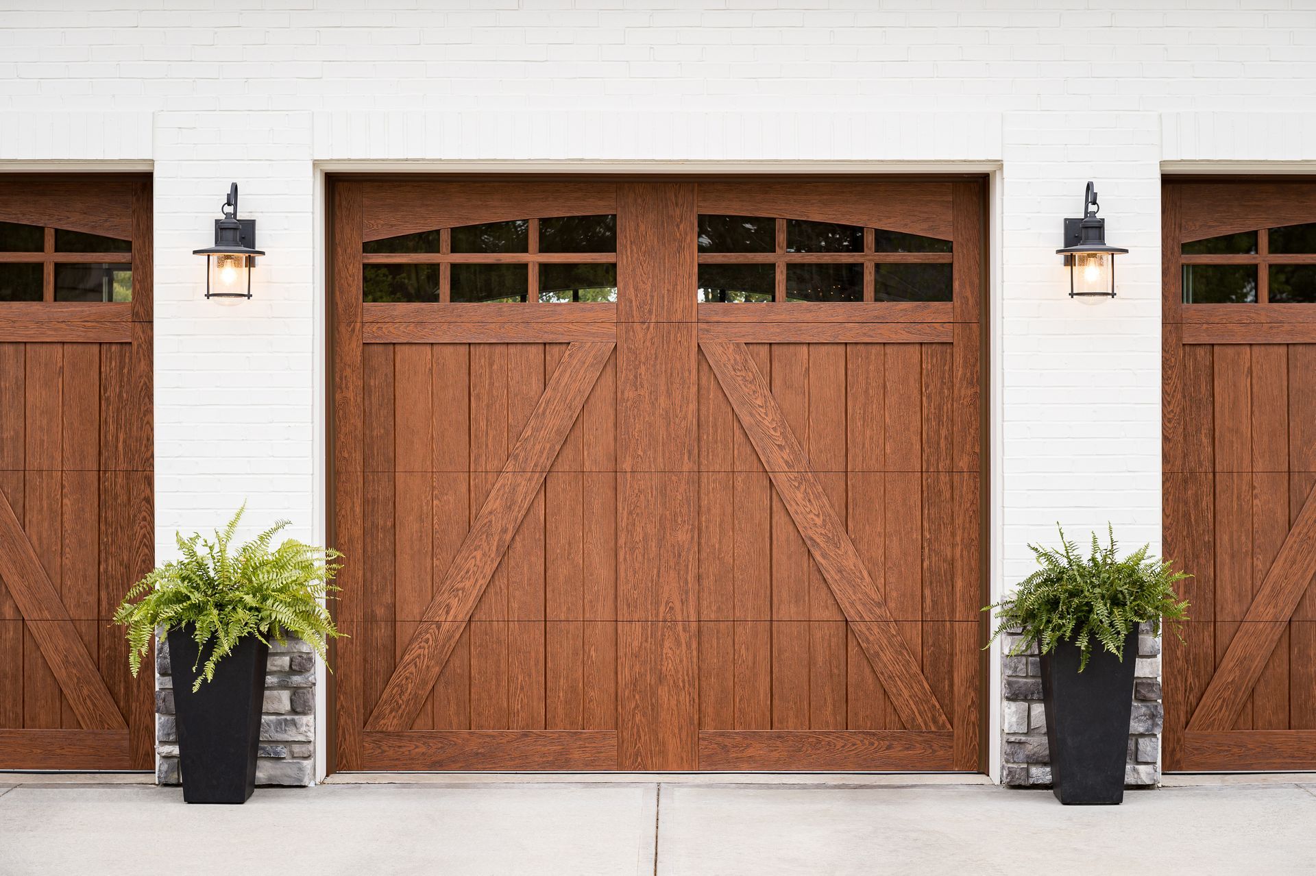 Three brown wooden garage doors with black lantern lights, white trim, and black planters with ferns.