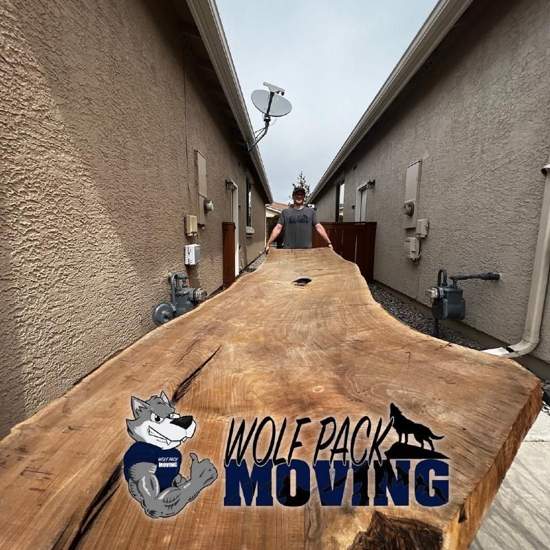 Man carries a large wooden table through a narrow space between two buildings. Wolf Pack Moving logo.