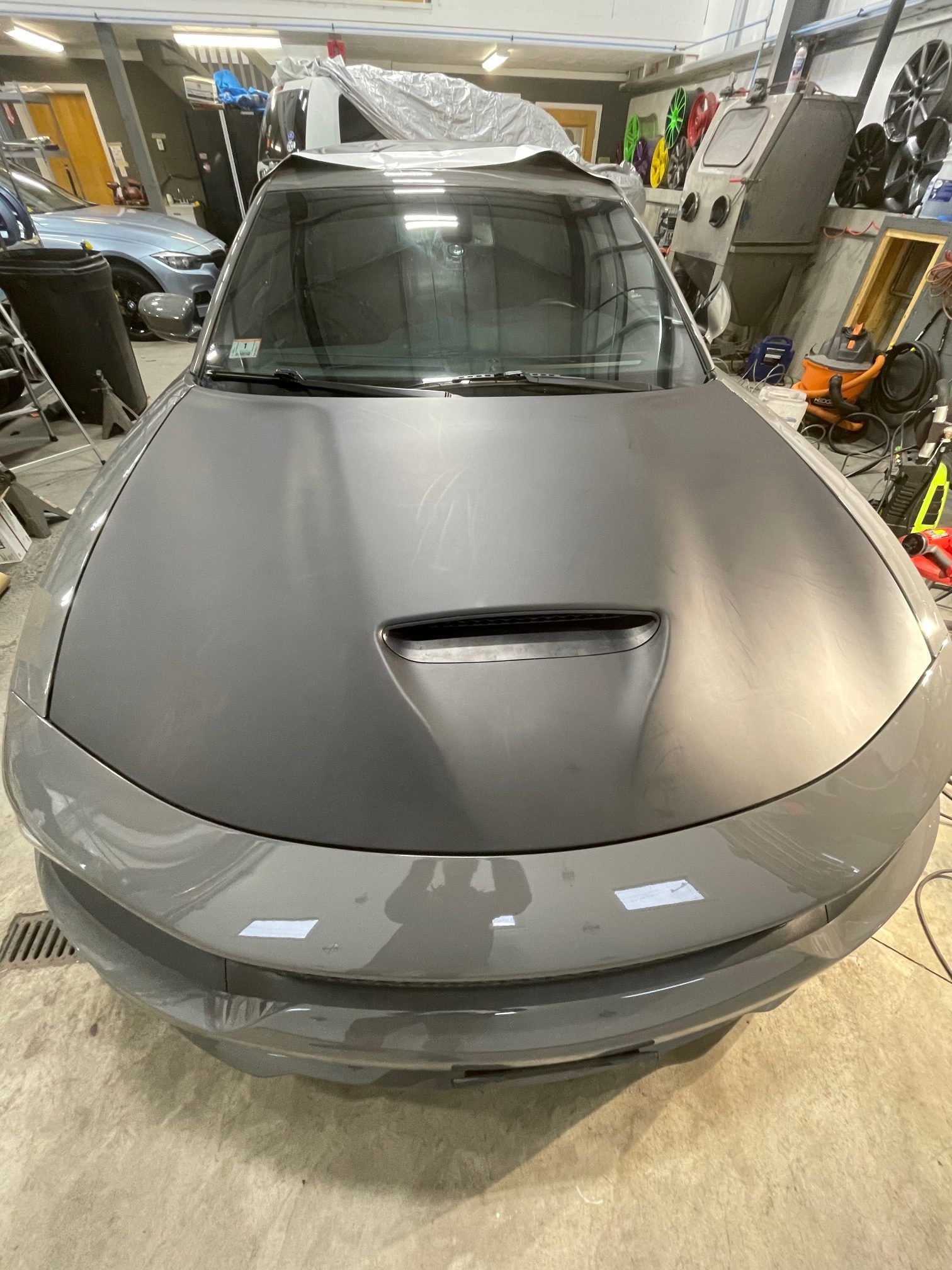 A gray car with a black hood is parked in a garage.