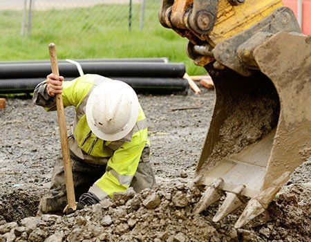 A construction worker in a high-visibility jacket and hard hat digs in the dirt near the bucket of an excavator.