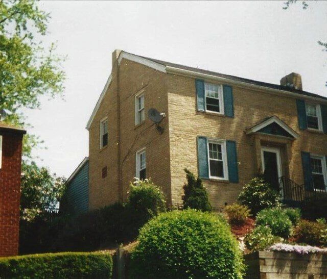 A two-story tan brick house with blue shutters, nestled on a small hill with lush green landscaping in the foreground.