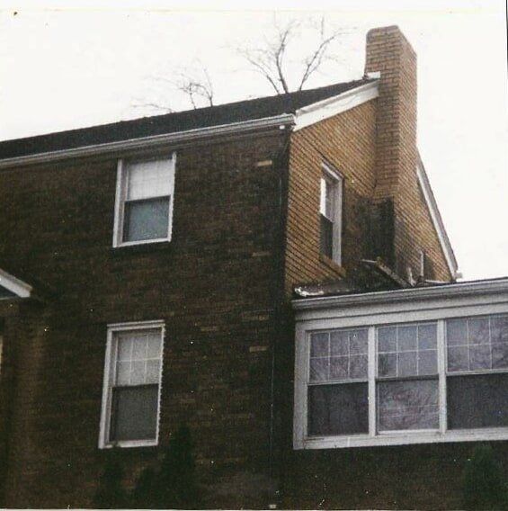 The exterior of a two-story brown brick house with a tall chimney, a white-framed windowed addition, and dark roof tiles.